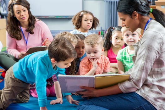 Female teacher giving a lesson to nursery students. They are sitting on the floor and there is a teacher taking notes.