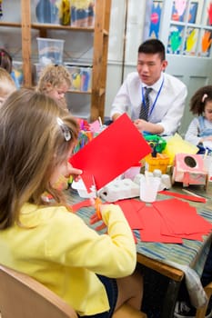 Little girl using scissors to cut shapes from a red piece of paper. She is in a nursery class with other students and a teacher.