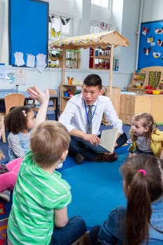 children are being read a story in nursery by a male teacher. One child has his hand up and the teacher is talking to him.