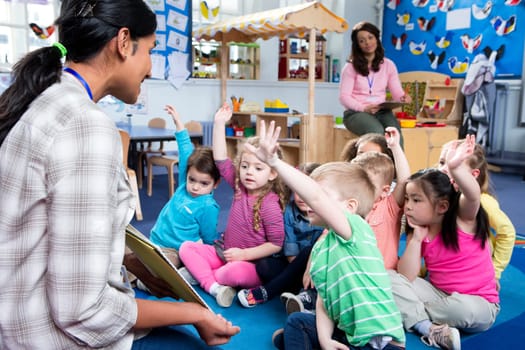 Female teacher giving a lesson to nursery students. They are sitting on the floor and some have their hands up to ask a question.