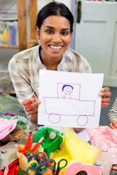 Nursery teacher proudly holding up her drawing to the camera. she is sitting in a classroom.