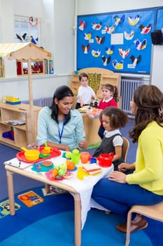 Teachers playing with plastic kitchen toys with their nursery students in the classroom.