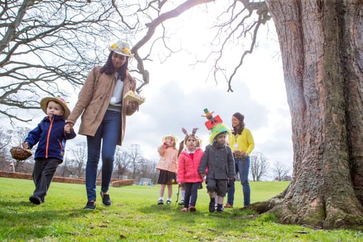 Nursery teachers and students doing an Easter egg hunt outdoors. They are wearing handmade hats and carrying baskets.