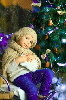 the girl in lilac jeans, in a knitted cap and with a knitted scarf sits on a sledge near a Christmas fir-tree with a toy and it is snowing