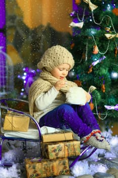 the girl in lilac jeans, in a knitted cap and with a knitted scarf sits on a sledge near a Christmas fir-tree with a toy and it is snowing