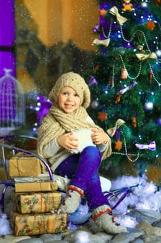 the girl in lilac jeans, in a knitted cap and with a knitted scarf sits on a sledge near a Christmas fir-tree with a toy and it is snowing