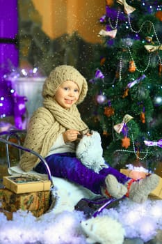 the girl in lilac jeans, in a knitted cap and with a knitted scarf sits on a sledge near a Christmas fir-tree with a toy and it is snowing