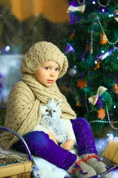 the girl in lilac jeans, in a knitted cap and with a knitted scarf sits on a sledge near a Christmas fir-tree with a toy and it is snowing