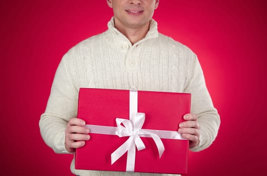 Young man in woolen sweater showing red giftbox with white ribbon