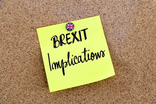 Yellow paper note pinned on cork board with Great Britain flag thumbtack, written text BREXIT IMPLICATIONS, United Kingdom exit from European Union