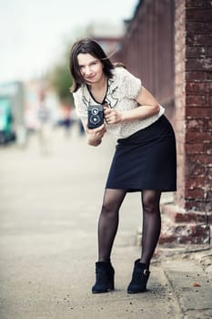 Girl holding retro camera and taking photo. Pretty young woman photographer with old vintage camera outdoors. Retro style photo. Selective focus on model. Vertical shot. Vintage style photo.