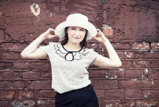 Pretty young woman in white blouse and hat posing against brick wall background. Woman holds the edges of the hat by hands. Toned photo with copy space. Vintage style photo.