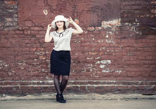 Pretty young woman in white hat, blouse and black skirt, standing against brick wall background. Girl holds the edges of the hat by hands. Toned photo with copy space. Vintage style photo.