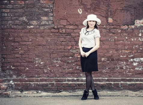 Pretty young woman in a white hat, blouse and black skirt, standing against old vintage brick wall background. Vintage portrait of a woman. Toned photo with copy space. Vintage style photo.