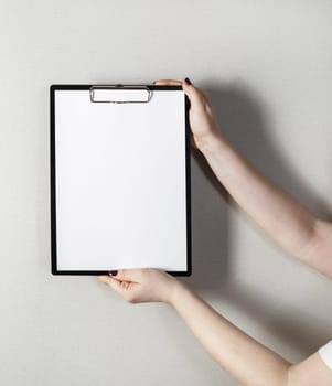 Clipboard with a blank sheet of paper in female hands with a deep shadow on the background of gray wall. Mock-up for design presentations and portfolios.
