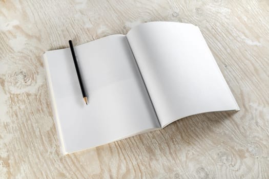 Blank opened brochure with a pencil on light wooden background with soft shadows. Template for design presentations and portfolios. Top view.