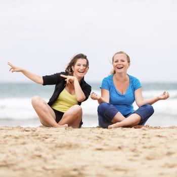 Two beautiful laughing young women have fun and enjoy sitting on the sand at the beach. Shallow depth of field. Focus on the models.