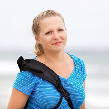 Photo of a smiling young blonde woman in a blue t-shirt with a backpack strap over his shoulder outdoors. Shallow depth of field. Focus on the model's face.