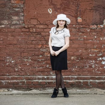 Photo of a woman in a white hat, blouse and black skirt, standing against the backdrop of an old vintage brown brick wall. Space for text.