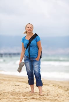 Young blonde woman barefoot in blue t-shirt and jeans with a backpack standing on the beach against the sea.  Shallow depth of field. Focus on model. Vertical shot.