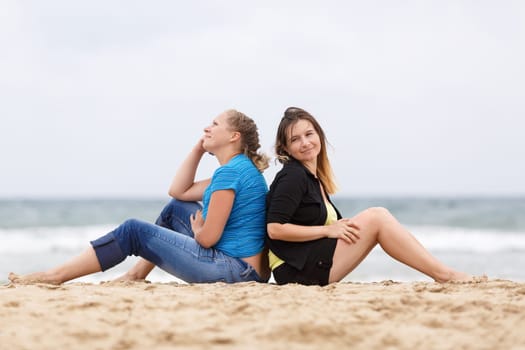 Two pretty young smiling woman sitting and relaxing on the sand on the seashore. Shallow depth of field. Focus on models.