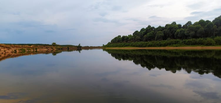Bend of the river. Evening summer landscape. Reflection shores and bushes in calm water. Panoramic shot.