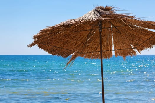 Beach straw umbrella against the bright blue sky and a calm sea. Selective focus on the parasol.