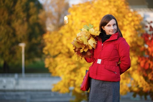 Female portrait against a bright autumn foliage. Young woman in red jacket holding in your hand a yellow maple leaves. Selective focus on model.