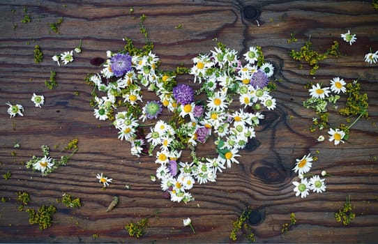 Heart symbol made of wild flowers and leaves on vintage wooden table background. Top view.