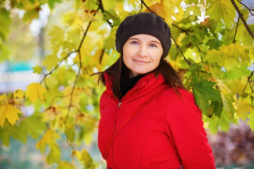 Pretty young woman in a beret and red jacket on the background of green and yellow autumn maple leaves. Female portrait. Selective focus on model.