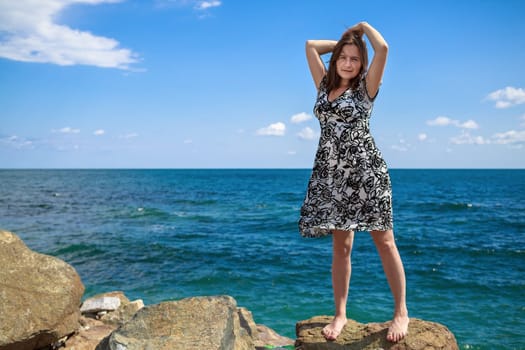 Pretty young woman standing on a stone and posing with his hands behind his head against a background of sea and blue sky. Selective focus on the model.