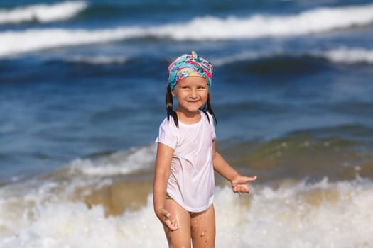 Happy child in a white t-shirt and colorful headdress against a blurred background of ocean waves on a sunny day. Shallow depth of field. Focus on the model's face.