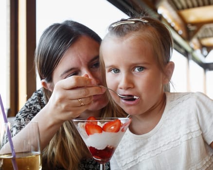 Mother feeds his daughter delicious sweet ice cream with strawberries in a restaurant. Shallow depth of field. Focus on the child's face.