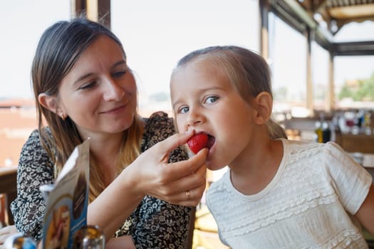Mum feeds the child delicious sweet strawberries in the restaurant. Shallow depth of field. Focus on the child's face.