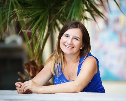 Pretty young smiling woman in a blue t-shirt sitting at a table outdoors on the background of a palm tree. Shallow depth of field. Selective focus on the model's face.
