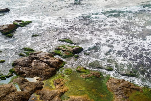 Close-up of sea surf. Shore with sea water and large rocks covered with algae. Rocky coastline with sea water and boulders.