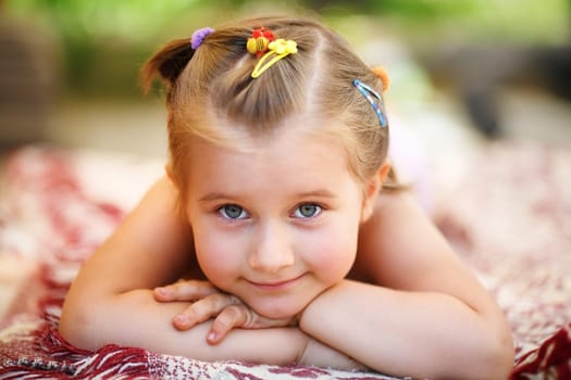 Sweet happy little baby girl lying on a blanket in the park. Close-up of child resting her head on her hands and looking into the camera. Shallow depth of field. Selective focus.