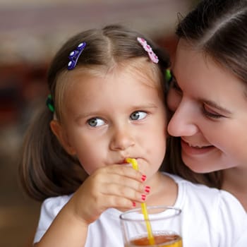 Closeup facial portrait of a little girl drinking juice through a straw next to her mother. Shallow depth of field. Focus on the model's face.
