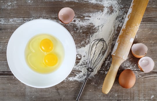 Cooking background with eggs, raw eggs in a dish, eggshells, flour, rolling pin and whisk on a wooden background. Top view.