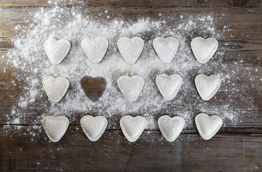 Raw ravioli in the shape of hearts, sprinkle with flour, on dark wooden background. Cooking dumplings. Top view.