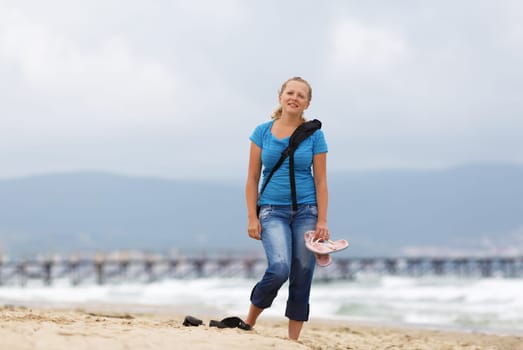 Young smiling blonde woman barefoot in blue t-shirt and jeans with a backpack standing on the beach against the sea.  Shallow depth of field. Focus on model.