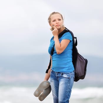 Pretty young blonde woman in a blue t-shirt and jeans with a backpack standing on a sea background.  Shallow depth of field. Focus on model.