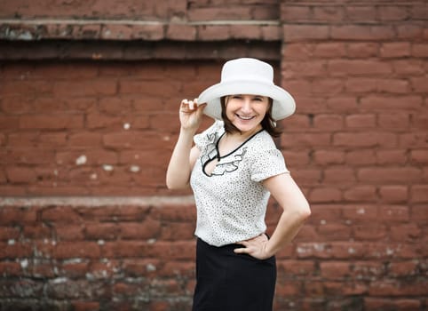 A smiling young woman in a white blouse and a hat posing against the backdrop of an old vintage brown brick wall. Female hand holds a hat.  Photo in retro style. Selective focus on model.