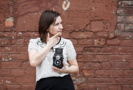 Pretty young woman photographer with retro camera in hand. Model is thinking and looking away. Old brown vintage grunge wall in the background.
