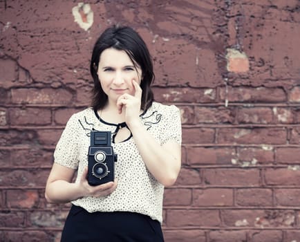 Woman photographer with vintage camera in hand on vintage brick wall background. Toned photo with copy space. Vintage style photo.