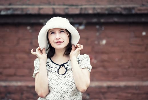 Outdoor lifestyle portrait of pretty young woman in white hat on brick wall background. Girl posing and showing tongue. Vintage style photo. Toned photo with copy space.