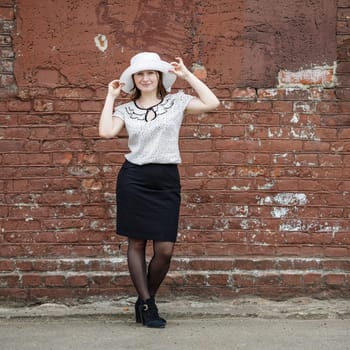 Photo of a woman in a white hat, blouse and black skirt, standing against the backdrop of an old vintage brown brick wall. Model holds the edges of the hat by hand. Portrait of a woman in a retro style.
