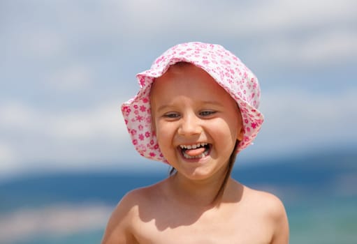 Adorable baby girl with a hat on his head on a blurred background of blue sky and sea. Laughing happy child. Hot sunny summer day. Selective focus.