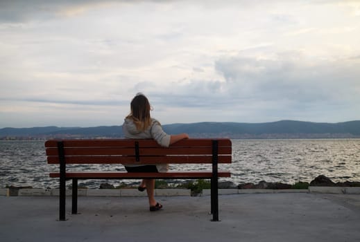 A woman sitting on a bench in the early morning and admire views of the bay and mountains. View from the back. Shallow depth of field. Focus on model.
