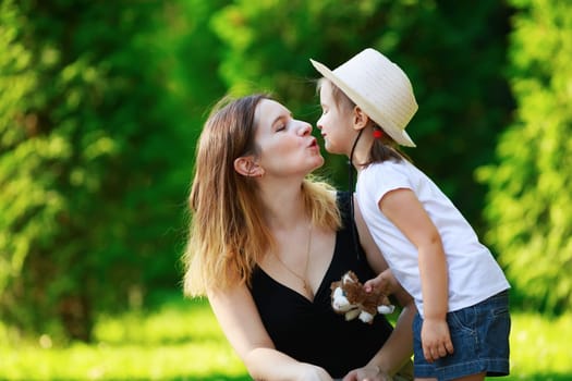 Mom kisses her little daughter. Mom and child girl. Happy family. Bright green foliage in the background. Sunny summer day. Shallow depth of field. Selective focus.